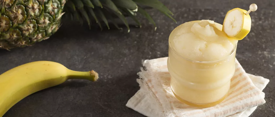 Glass of Banana ‘Nana Daiquiri garnished with  banana slice on white cloth alongside a banana