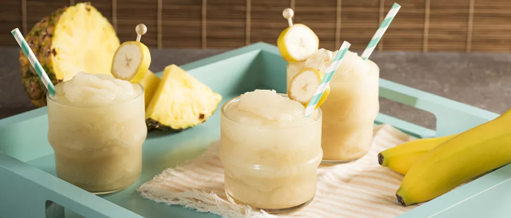 Three glasses of Banana ‘Nana Daiquiri garnished with straws and banana slice on blue tray alongside two bananas