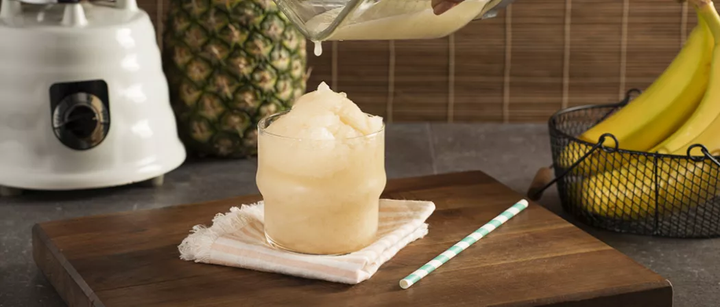 Glass of Banana ‘Nana Daiquiri on striped cloth coaster alongside a straws on wooden board