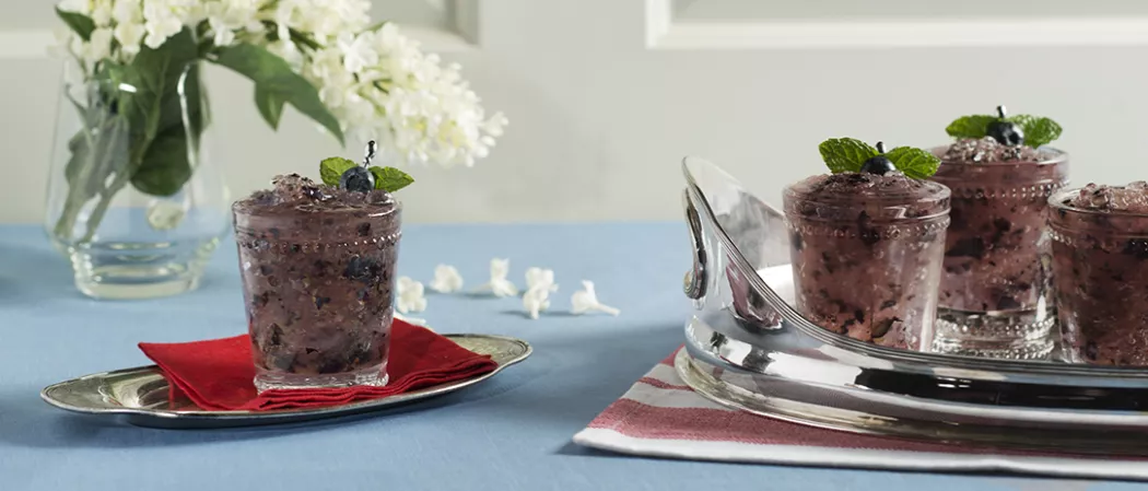 Glass of Blue Stallion Julep garnished with blueberry and mint leaves on red cloth on tray and three Glass of Blue Stallion Julep on another tray