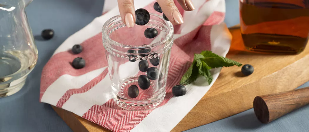 Perosn putting blueberries into empty glass alongside few blueberries and mint leaves 