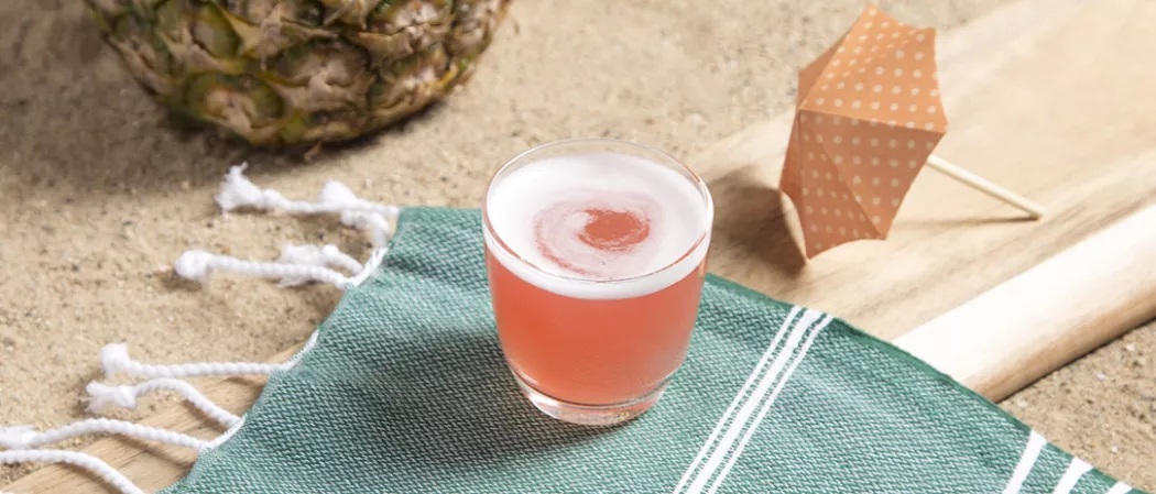 Glass of Sex On The Beach on green piece of cloth and paper parasol on wooden tray