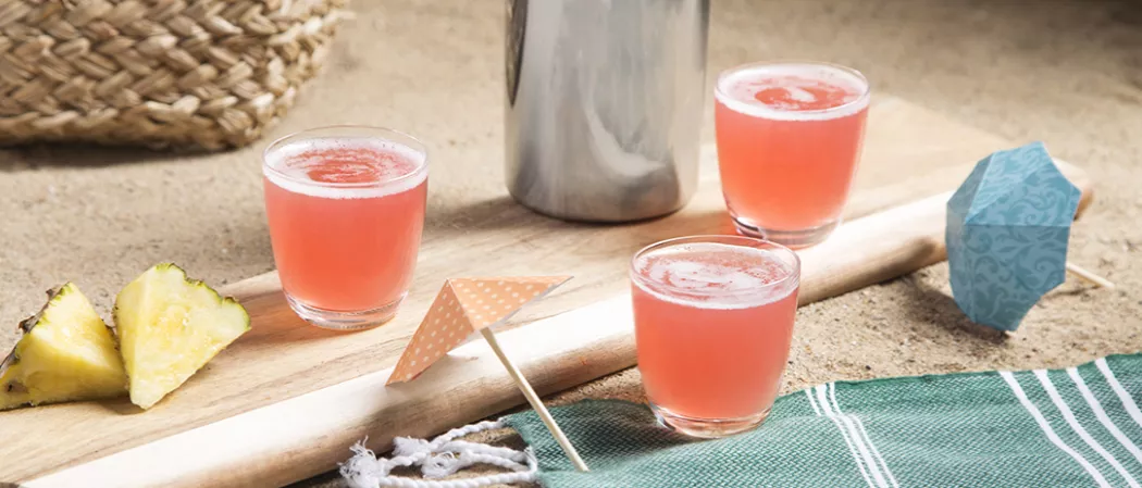 Two glasses of Sex On The Beach alongside pineapple wedges on wooden tray  alongside glass of Sex On The Beach and paper parasol