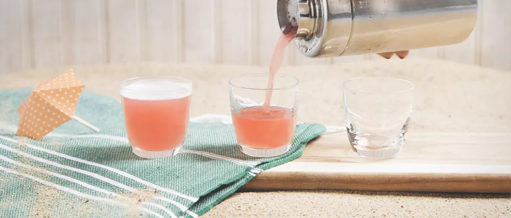 Person pouring Sex On The Beach into glass alongside a empty glass, a glass of Sex On The Beach and paper parasol