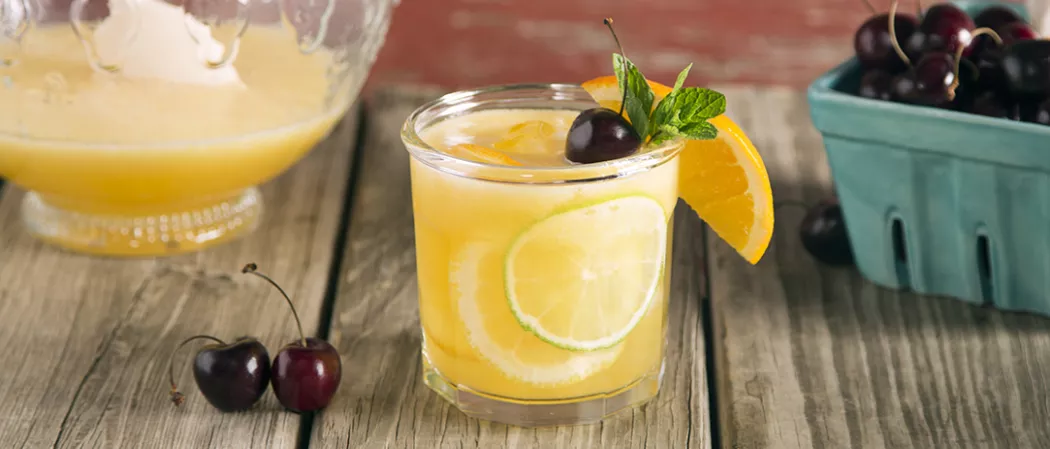 Glass of Sparkling Cherry Sangria garnished with orange wedge, lime slices, mint leaves and cherry alongside two cherries