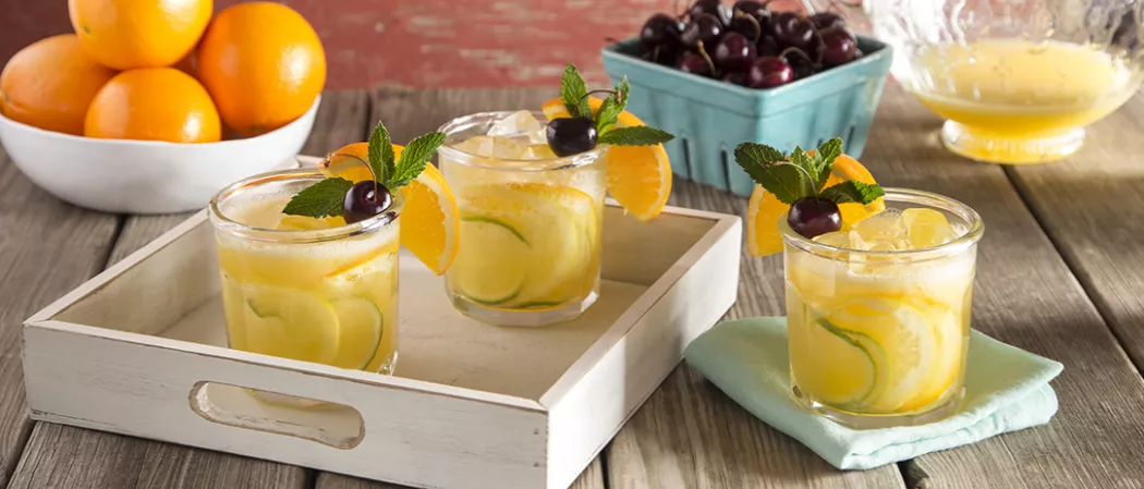 Two glasses of Sparkling Cherry Sangria garnished with orange wedge, lime slices, mint leaves and cherry on white tray alongside another glass of Sparkling Cherry Sangria on  blue cloth
