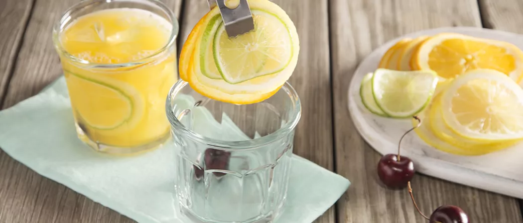 Person adding lime and lemon slices into empty glass alongside glass of Sparkling Cherry Sangria on blue piece of cloth alongside white plate with orange, lime and lemon slices