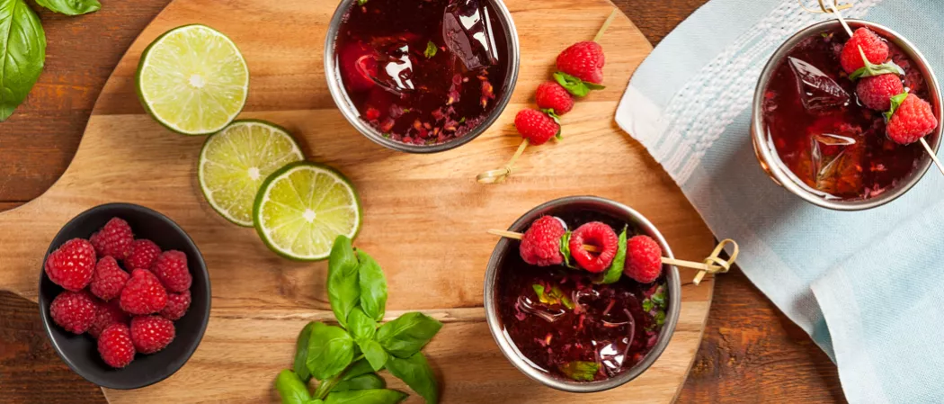 Three glasses of Berry Moscow Mule garnished with raspberries and mint leaves on wooden  board  alongside mint leaves, few lime slices and bowl of raspberries