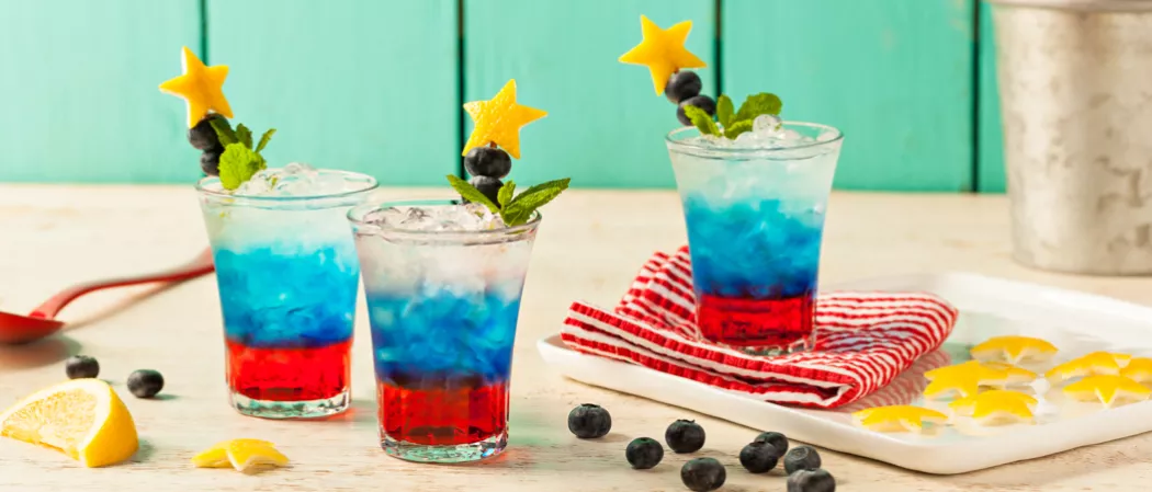 Three glasses of Citrus-Spangled Spritzer garnished with mint leaves, lemon rind star and berries one on red white striped cloth on white tray alongside some blueberries. lemon wedge and lemon rind star