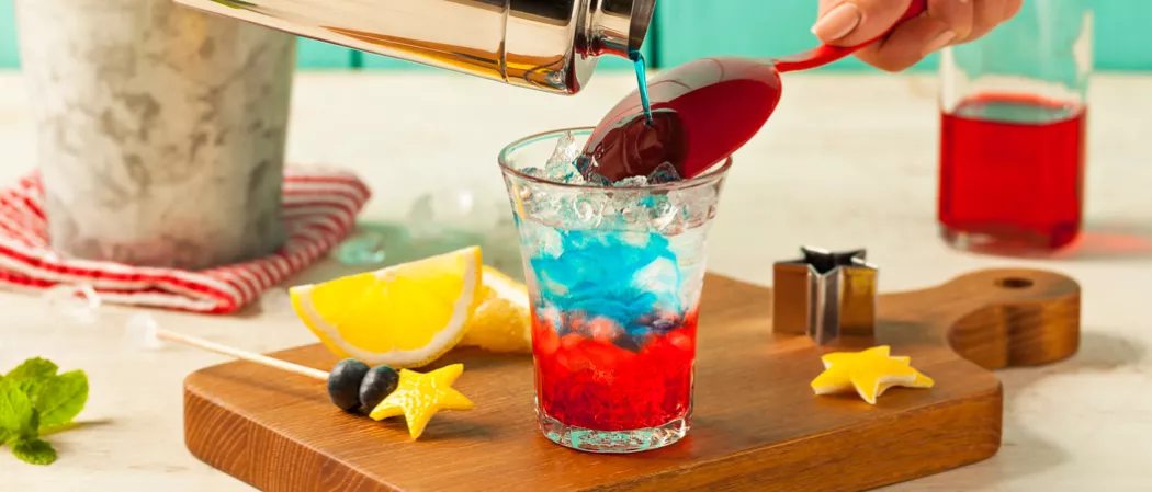 Person pouring Citrus-Spangled Spritzer into glass alongside lemon wedge, lemon rind star on wooden tray