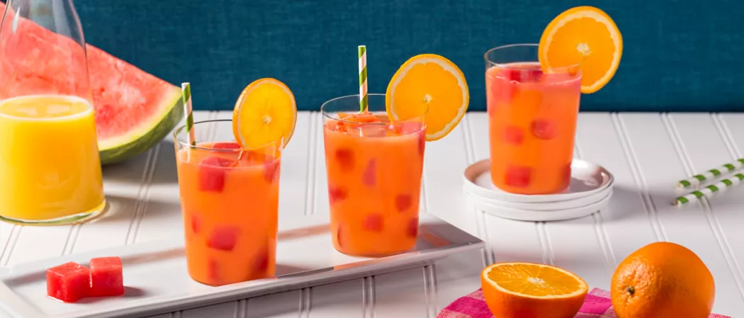 Three glasses of Juicy Screw garnished with orange slice and straws alongside half and whole orange and watermelon cube