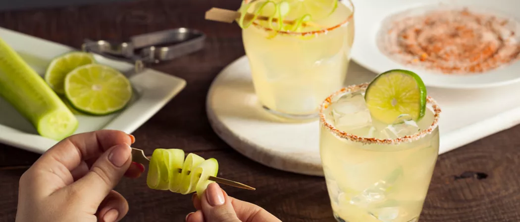 Person making thin cucumber slice garnish alongside two glasses of Spicy Cucumber Margarita garnished with lime slice