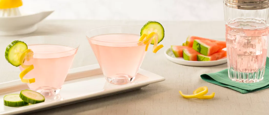 Two glasses of Cucumber Watermelon Martini, garnished with cucumber and lemon peel twists on the rim of the glasses, are arranged in a white tray. Alongside few lemon peel twists, a plate full of watermelon slices, and another glass of same