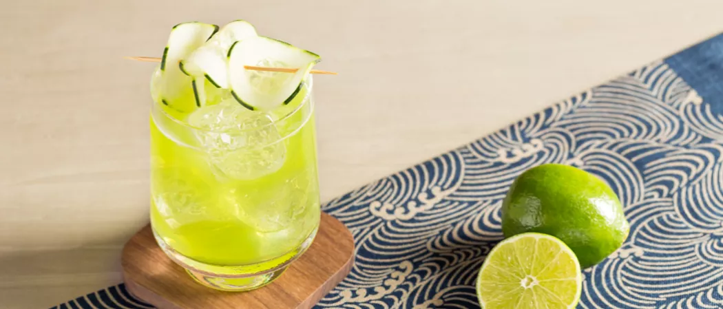 Glass of Cucumber Cooler garnished with cucumber slices on coaster alongside some limes