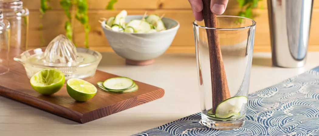 Person muddling cucumber slices into glass alongside a wooden tray with limes, cucumber slices and lemon juicer