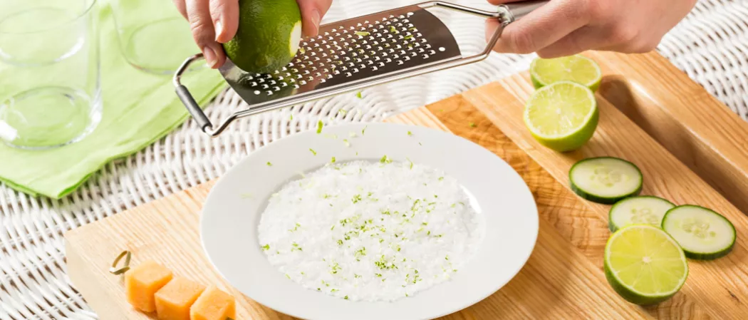 Person grating lime zest into plate alongside few melon cubes, few cucumber and lime slices on wooden board