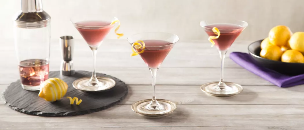Three glasses of Purple Haze Martini  one on tray with piled lemon, shaker, and jigger, two on table garnished with twisted lemon peel, bowl of lemons on purple cloth."