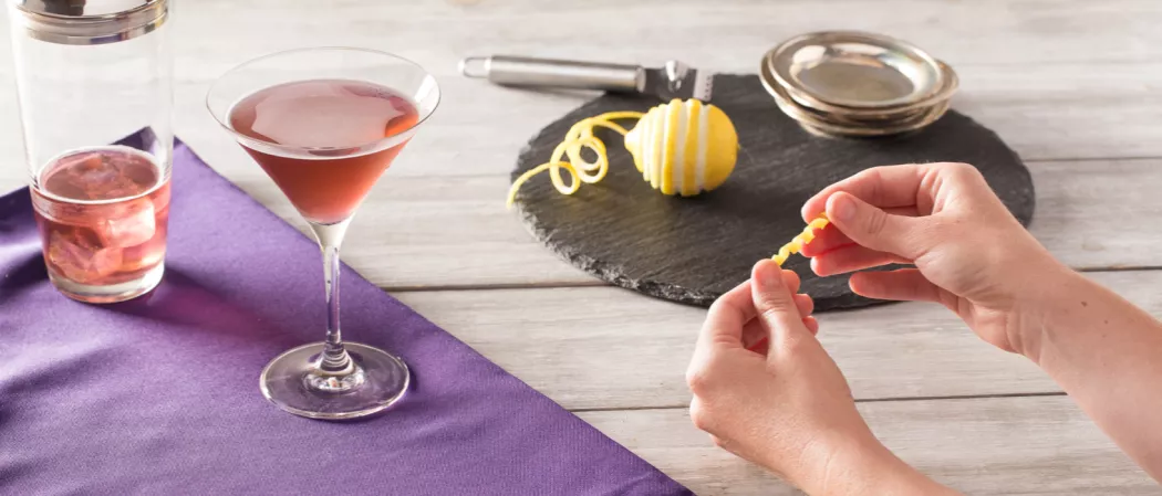 A person holding a lemon twist peel.alongside a  glass, shaker of Purple Haze Martini on a purple cloth, lemon twisted peel, shaker cap, and grater on a wooden coaster,