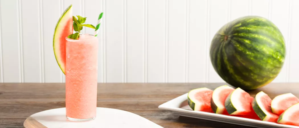 Glass of Downward Facing Daiquiri garnished with watermelon slice, mint leaves and a straws alongisde tray with watermelon slices and a watermelon