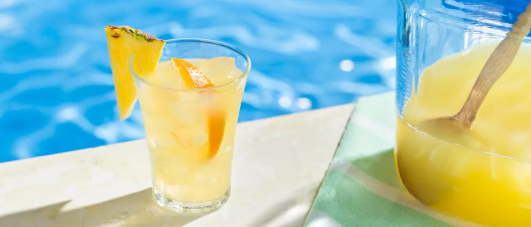 A glass of Endless Summer Punch garnished with ice cubes, an orange slice, and a pineapple slice on the rim, alongside a jar of Endless Summer Punch on a green cloth.