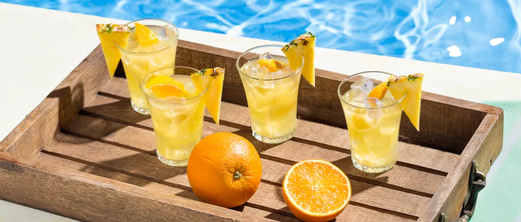 four glasses of Endless Summer Punch garnished with ice cubes, a slice of orange and a slice of pineapple on the rim of the glass. alongside an orange and a slice of orange, all in a wooden tray.