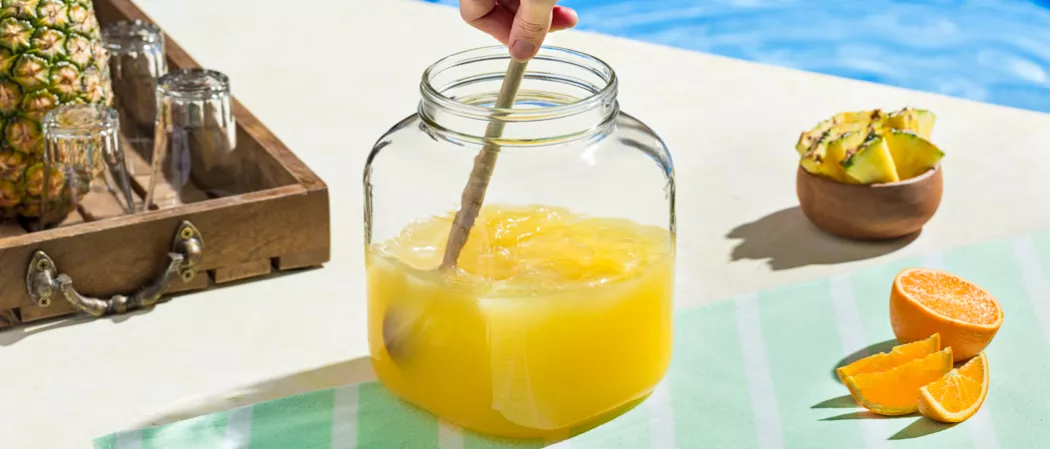 A person is stirring a jar of Endless Summer Punch alongside slices of orange, a bowl of pineapple and three inverted glasses and pineapples in a wooden tray.
