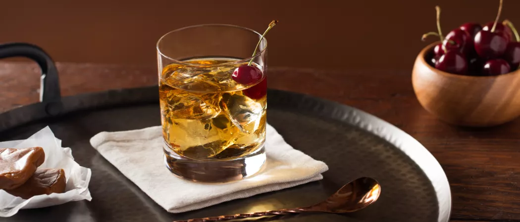A Buttershots® Old Fashioned with a cherry, served on white cloth beside a bar spoon, all on an iron plate with a bowl of cherries.