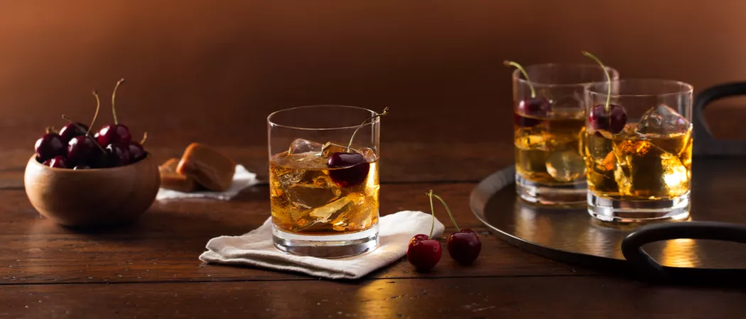Three glasses of Buttershots® Old Fashioned garnished with cherry one on a white cloth and two on a iron plate alongside  