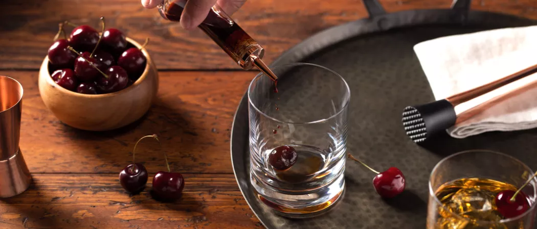 A person pours brown sugar syrup into a glass with a cherry, next to another glass of Buttershots® Old Fashioned with a cherry garnish on a white cloth and a crusher, all on an iron plate with a bowl of cherries nearby