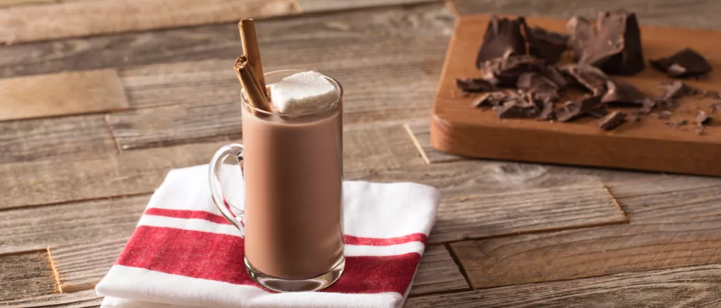 Fireside Coco in a mug on a napkin, topped with marshmallows and cinnamon sticks, next to chopped chocolate on a board