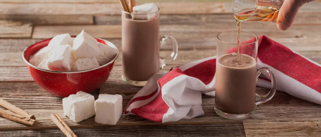Two glasses of Fireside Coco, one ready with Jim Beam Kentucky Fire, the other being poured. Nearby are a bowl of marshmallows , cinnamon sticks, and a napkin