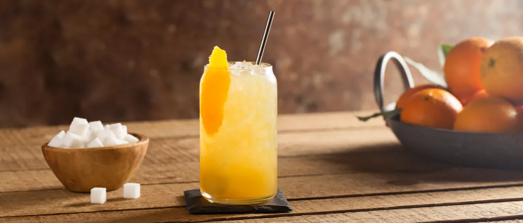  Glass of The Rider with ice and straws garnished with orange peel on black coaster alongside a bowl of suger and bakset of oranges