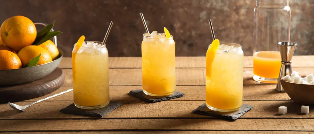 Three glasses of The Rider with ice and straws garnished with orange peel on black coaster alongside a bowl of suger, bakset of oranges, a jigger and a bar spoon.