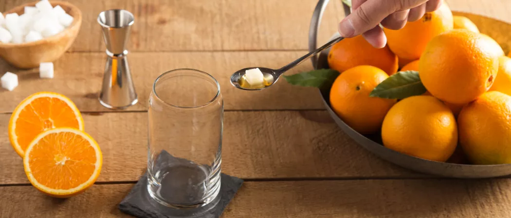 A person putting suger cube using spoon into empty glass on coaster alongside two orange slices, a jigger, bowl of sugar cubes and a basket of oranges