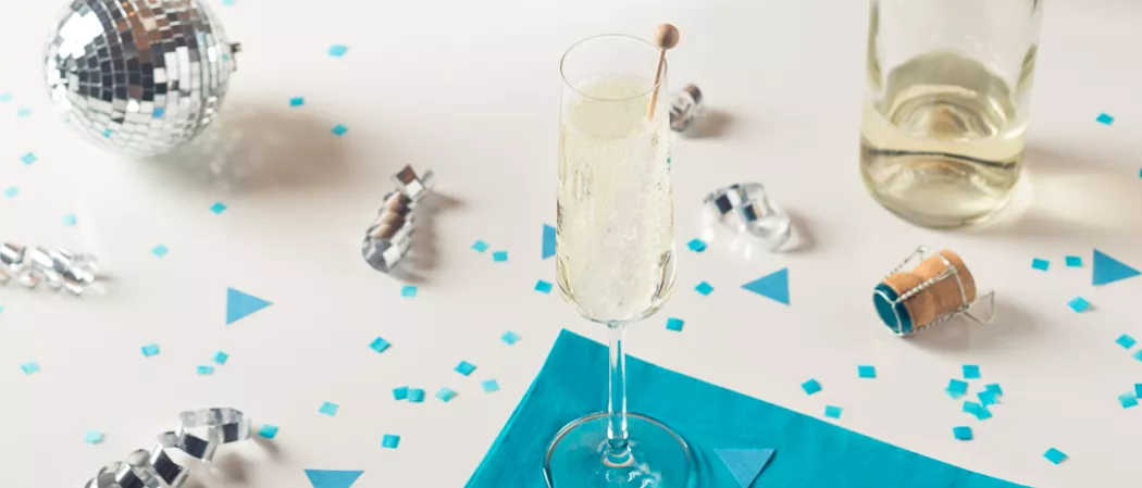 A flute glass of Midnight's Kiss, garnished with a rock candy stick, rests on a blue paper cloth, accompanied by small pieces of paper, a silver spiral sheet, disco ball, a bottle of Midnight's Kiss. 