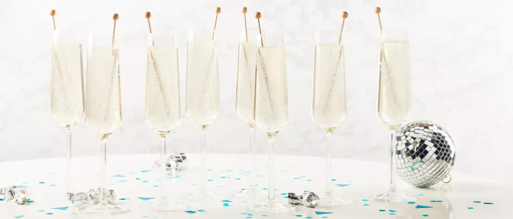 Eight glasses of Midnight's Kiss, each garnished with a rock candy stick, accompanied by a disco ball, small pieces of paper, and silver spiral sheets.