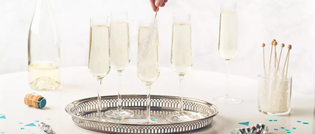 Five flute glasses of Midnight's Kiss, with four in a tray. One glass has a person stirring with a rock candy stick. Nearby, a bottle of the same drink with a fallen cap, a glass with rock candy sticks.