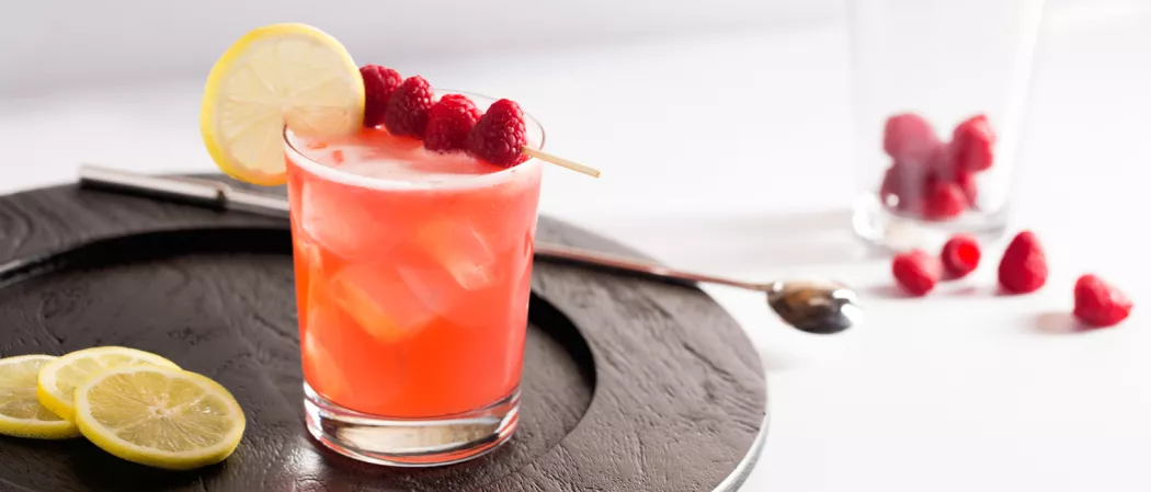 Glass of Ruby Sparkler garnished with lemon slice and raspberries alongside lemon slice on wooden tray