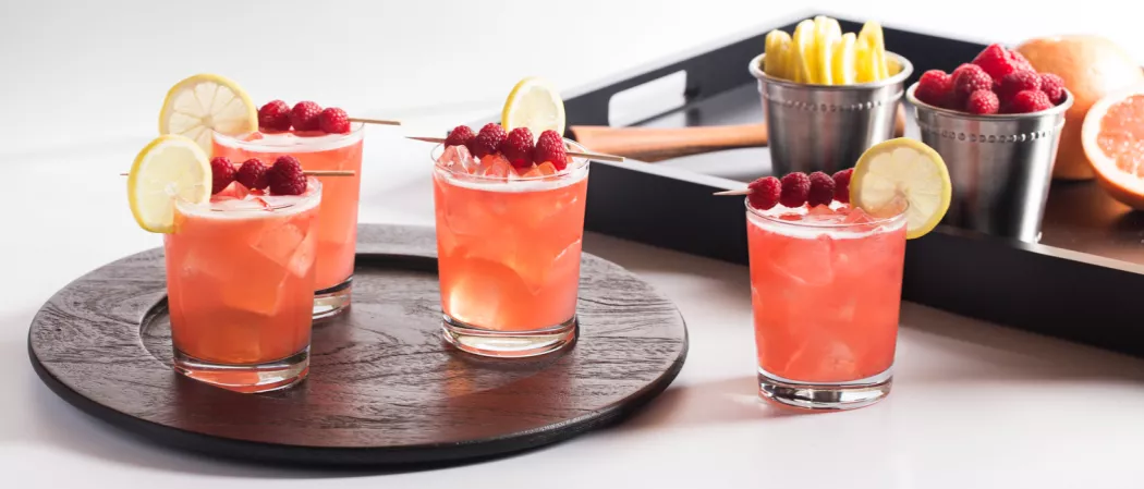 Three glasses of Ruby Sparkler garnished with lemon slice and raspberries on wooden tray alongside another glass of Ruby Sparkler 