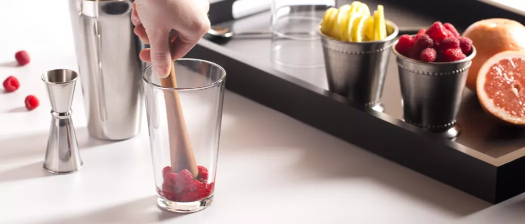 person muddling raspberries in glass alongside a jigger. some raspberries and a wooden tray with glasses of lemon slices and raspberries