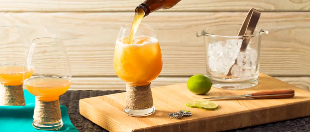person pouring Slowpoke Shandy into glass alongside bottle caps, lime slices, lime, knife and bucket of ice cubes