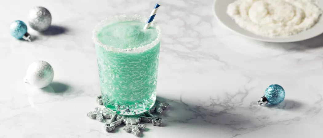 Glass of Blue Flurry rimmed with powdered sugar with straws on snowflake shaped coaster alongside few Christmas tree balls and plate of powdered sugar