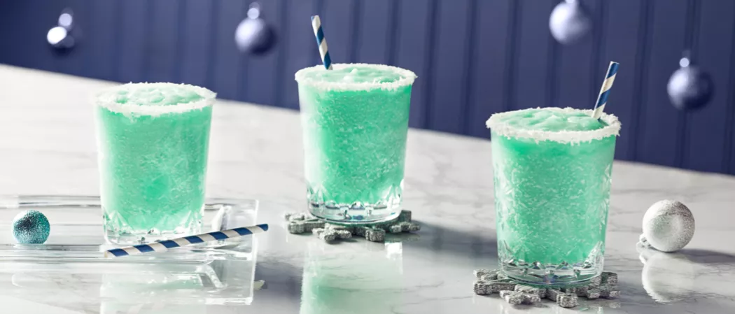 Two glasses of Blue Flurry rimmed with powdered sugar with straws on snowflake shaped coaster and another glass of Blue Flurry on transparent tray with a straws
