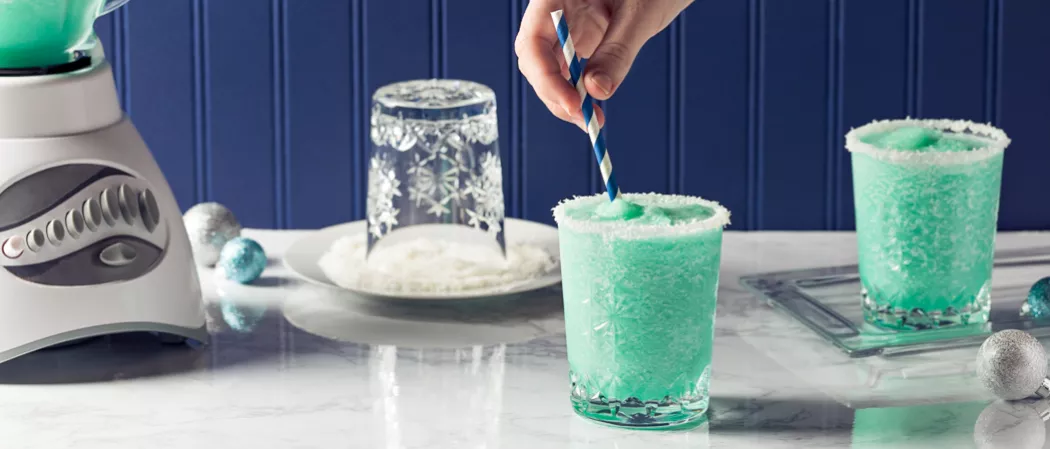 Person stirring glass Blue Flurry with straws alongside another glass of Blue Flurry rimmed with powdered sugar and a empty upside down glass on plate with powdered sugar