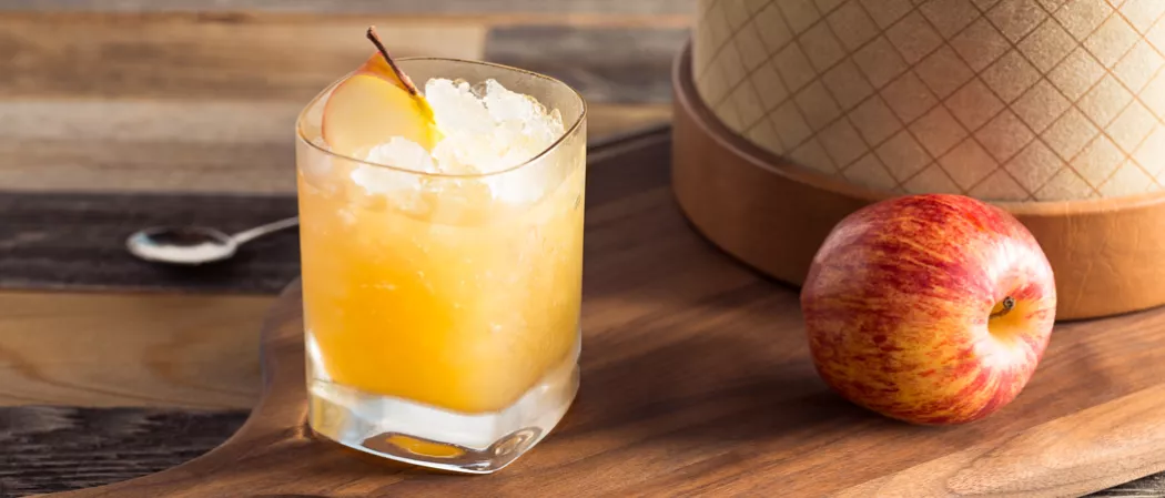 Glass of Bourbon Cider Sour garnished with a slice of apple and some crushed ice, next to an apple on a chopping board