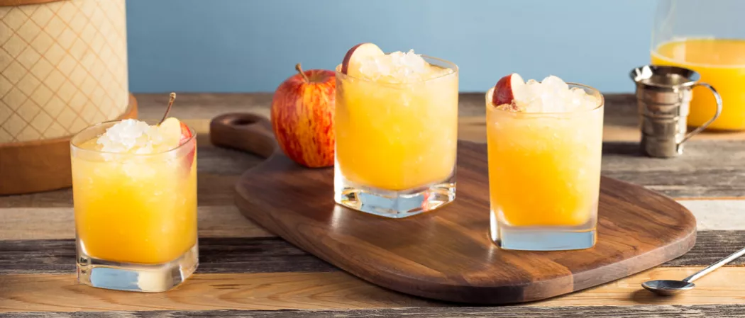 An apple, three glasses of Bourbon Cider Sours garnished with apple slices and ice, served on a board with a spoon near by
