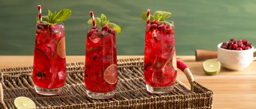 Three glasses of Cranberry Mojito with ice cubes, lime slice and cranberries and straws garnished with mint leaves and a lime slice on wooden tray alongside a bowl of cranberries, a lime slice and a muddle