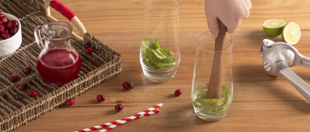 A person crushing mint leaves using muddle in a glass alongside another glass with mint leaves, few cranberries, two straws. a lime juicer, two lime slices and a tray with cranberries a jug of Cranberry Mojito and bowl of cranberries