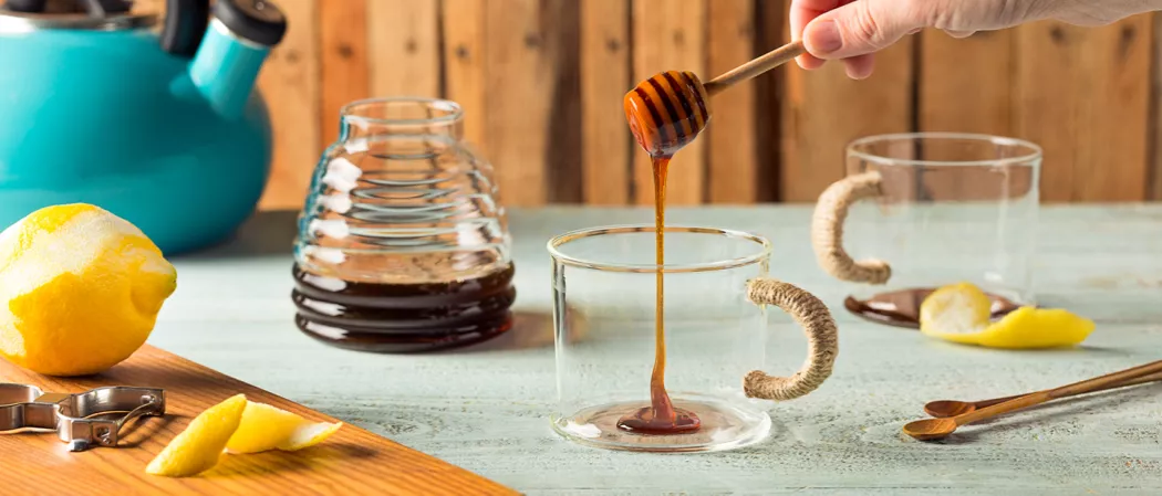 Person addnig honey into empty cup using honey dipper alongside some spoons, empty cup and honey pot