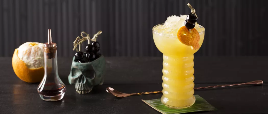 Glass of Zombie garnished with crushed ice, orange peel and cherries on coaster alongside bottle, cherries in skull shaped pot, a bar spoon and orange